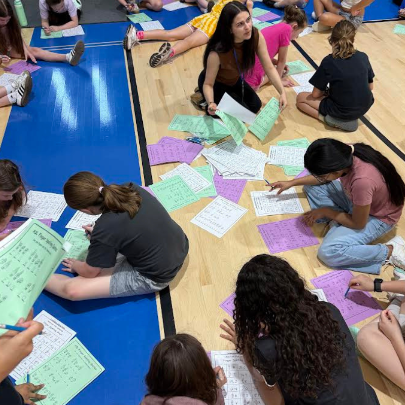 A group of students and an instructor work on worksheets spread across a gym floor.