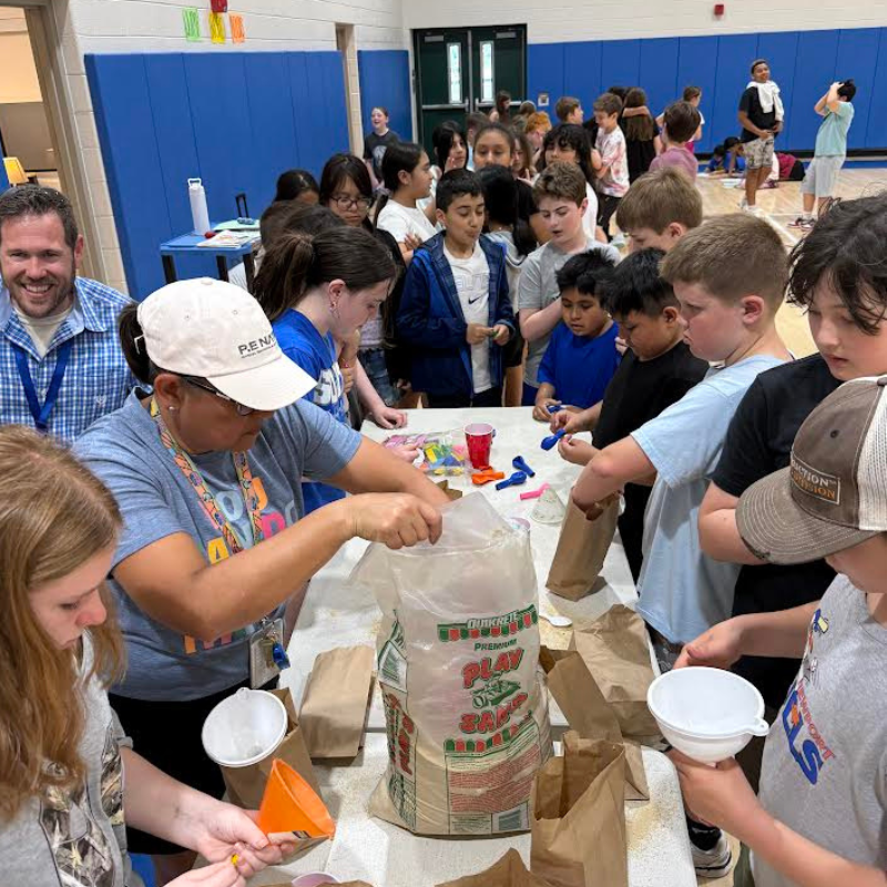 Children and adults gather around a table in a gymnasium, participating in a craft activity with sand and bags.