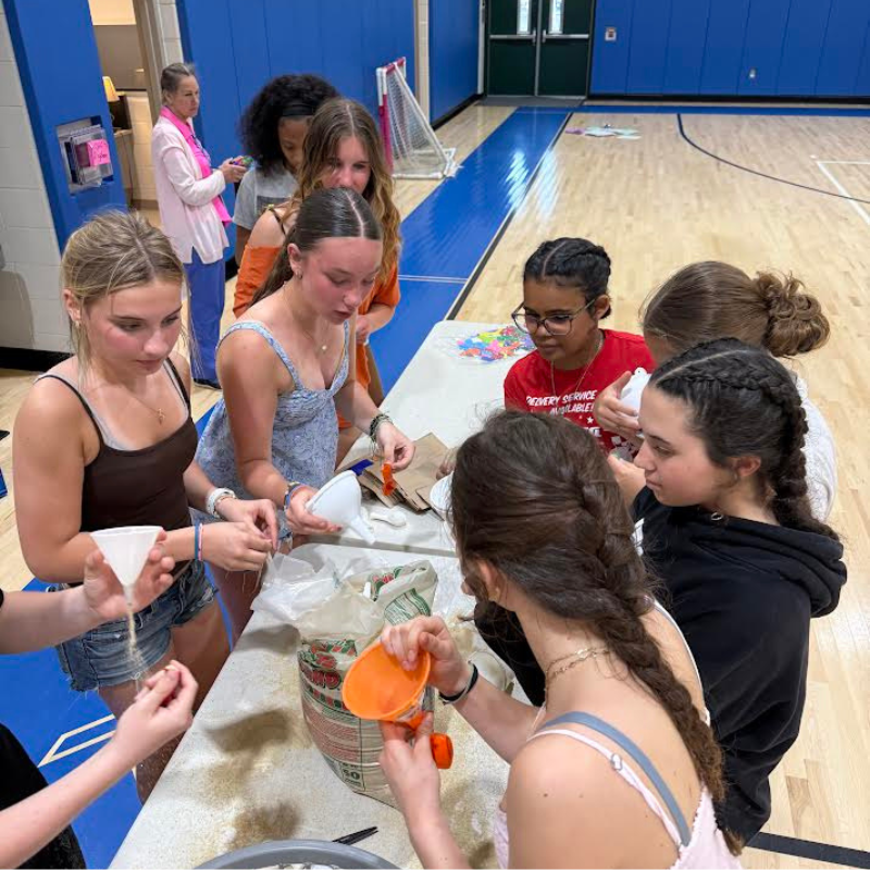 A group of young people are gathered around a table in a gymnasium, engaged in a craft activity involving sand and funnels.