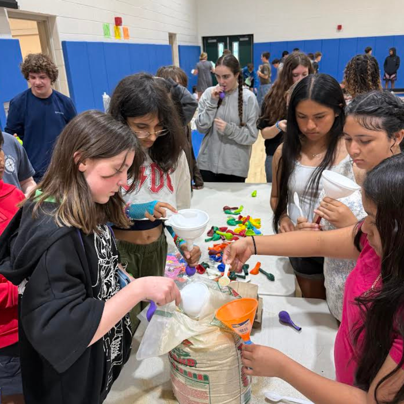 Young people fill balloons with a white substance using funnels and scoops at a table.