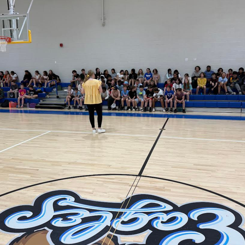 A woman in a yellow top stands on a basketball court facing an audience seated on bleachers.
