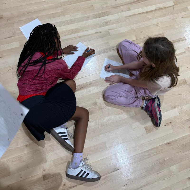 Two young girls sit on a wooden floor, writing on papers with pens.