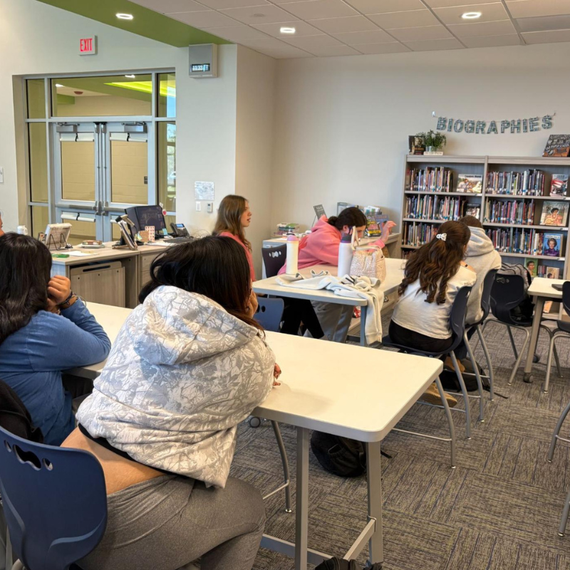Students sit at tables in a classroom or library, facing a bookshelf labeled 'BIOGRAPHIES'.