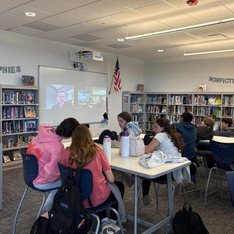 Students gather around a table in a library, watching a video conference on a projector screen.