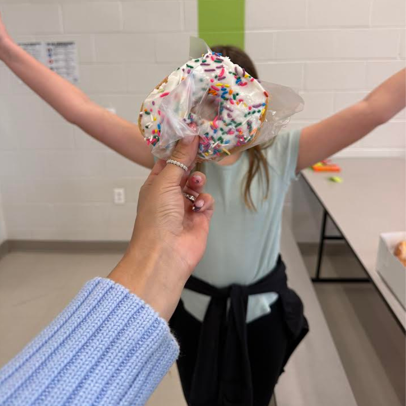 A person holds a frosted donut with colorful sprinkles towards the camera, with a child in the background.