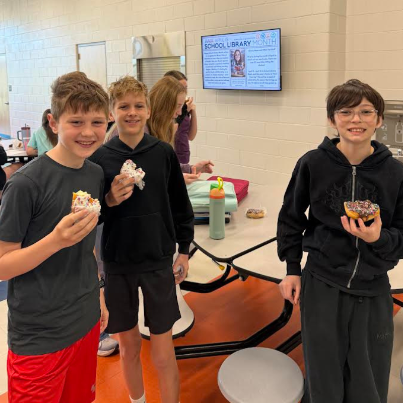Three smiling boys hold donuts in a school cafeteria, with a "School Library Month" display on the wall.
