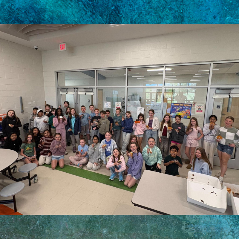 A diverse group of students poses for a photo while holding donuts in a school hallway.