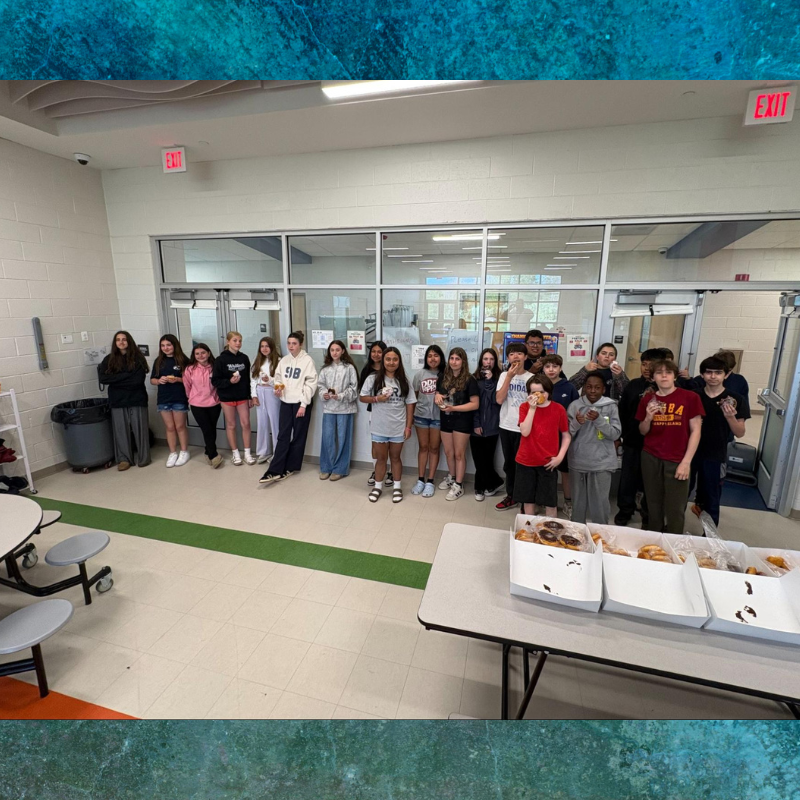 A group of students stands in a hallway, some holding donuts, with boxes of donuts on a table.