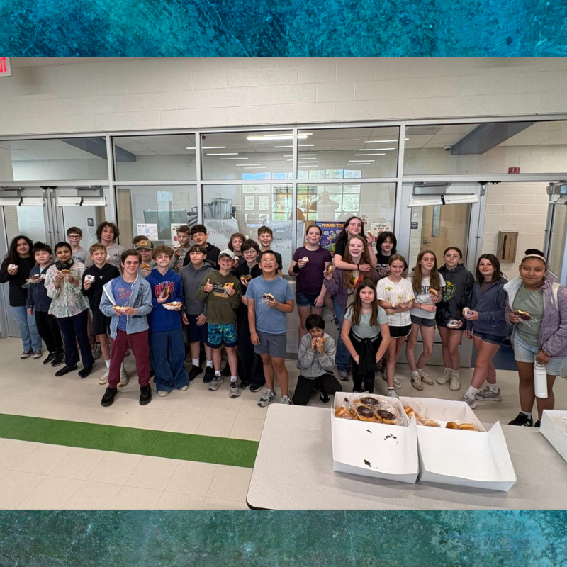 A diverse group of students stands together, many holding donuts, in a school hallway.