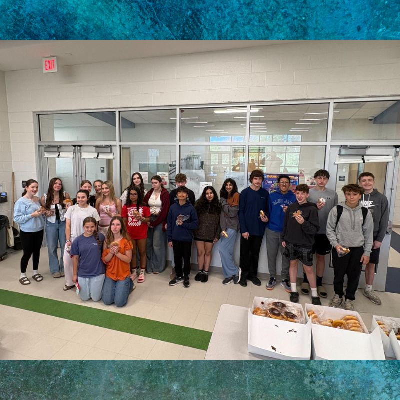 A group of teenagers stands together indoors, holding donuts and smiling.