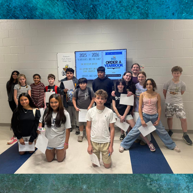 A group of middle school students stand in a hallway, holding papers, with a screen displaying yearbook information.