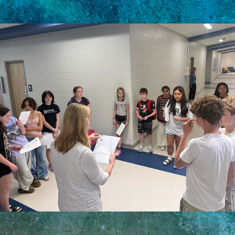 A teacher holds papers while a group of students listens attentively in a school hallway.