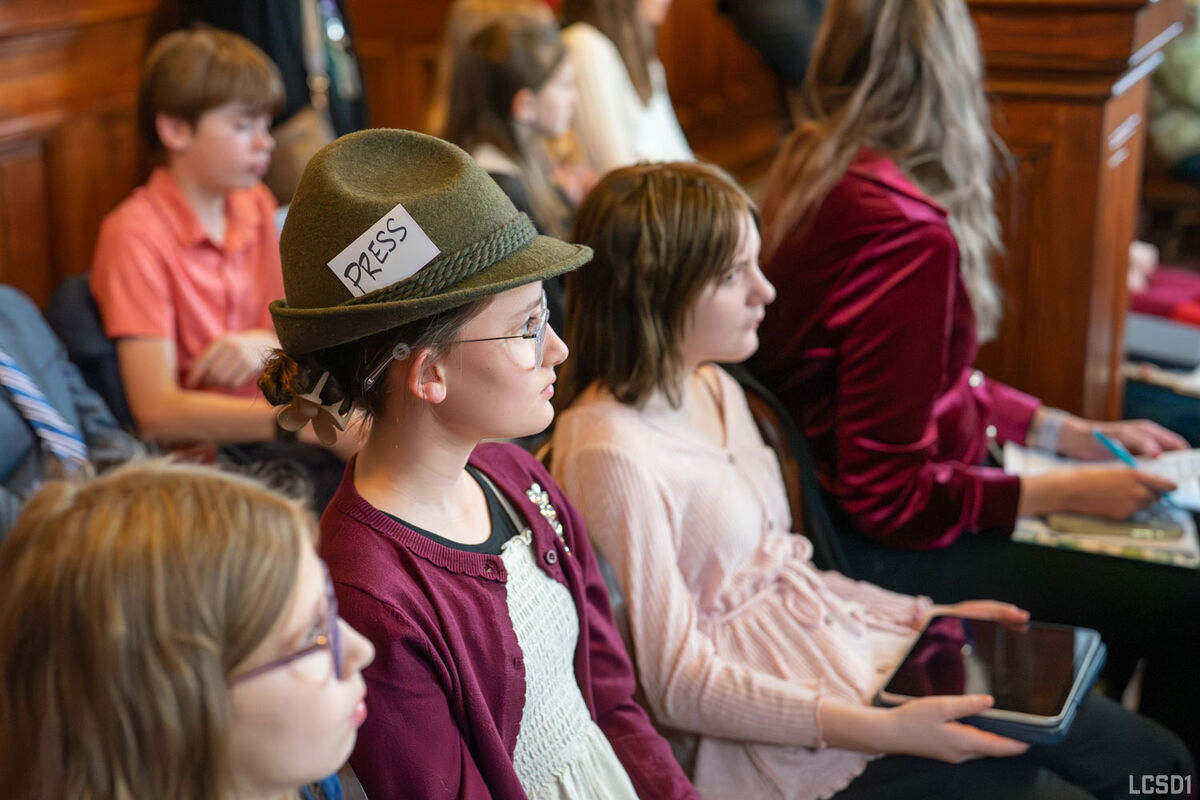 A student wearing a green cap with a tag that says "press" sits in a crowd.