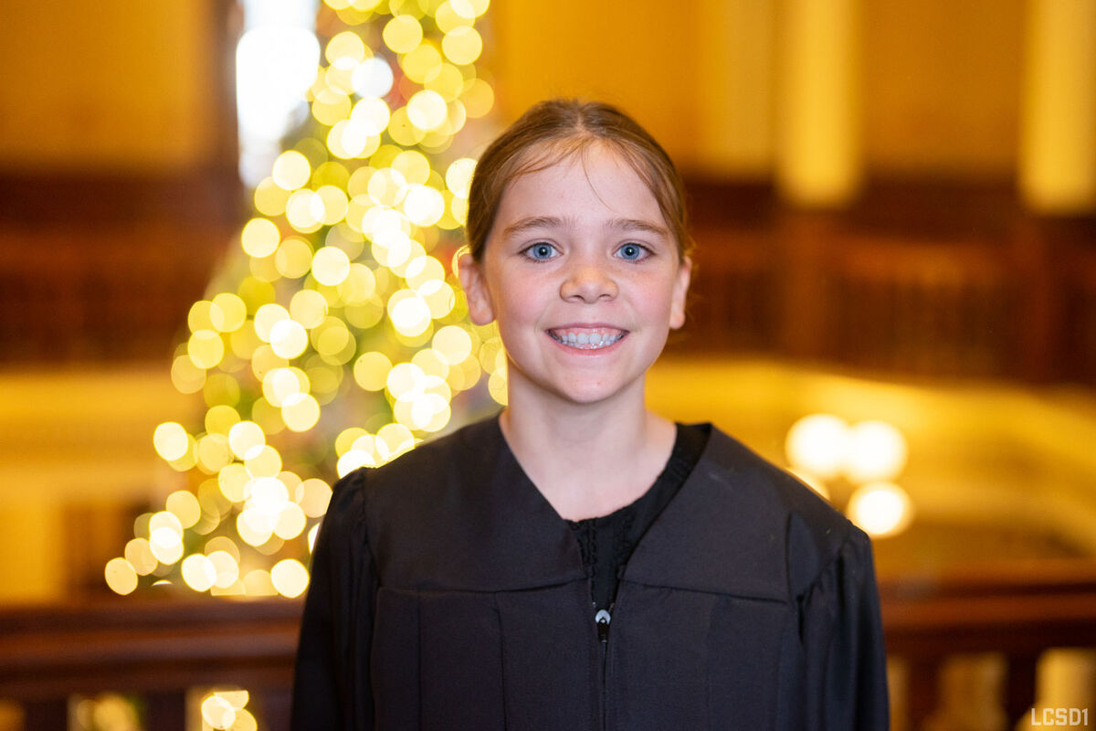 A student wearing a judge's gown smiles in front of a glowing Christmas tree