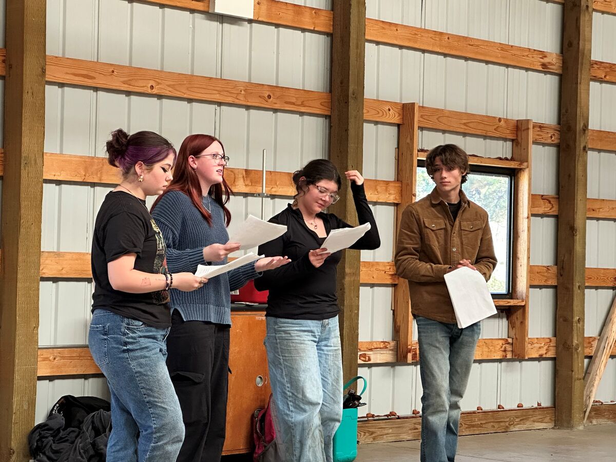Four young adults stand inside a rustic building with wooden beams, holding papers and rehearsing together.