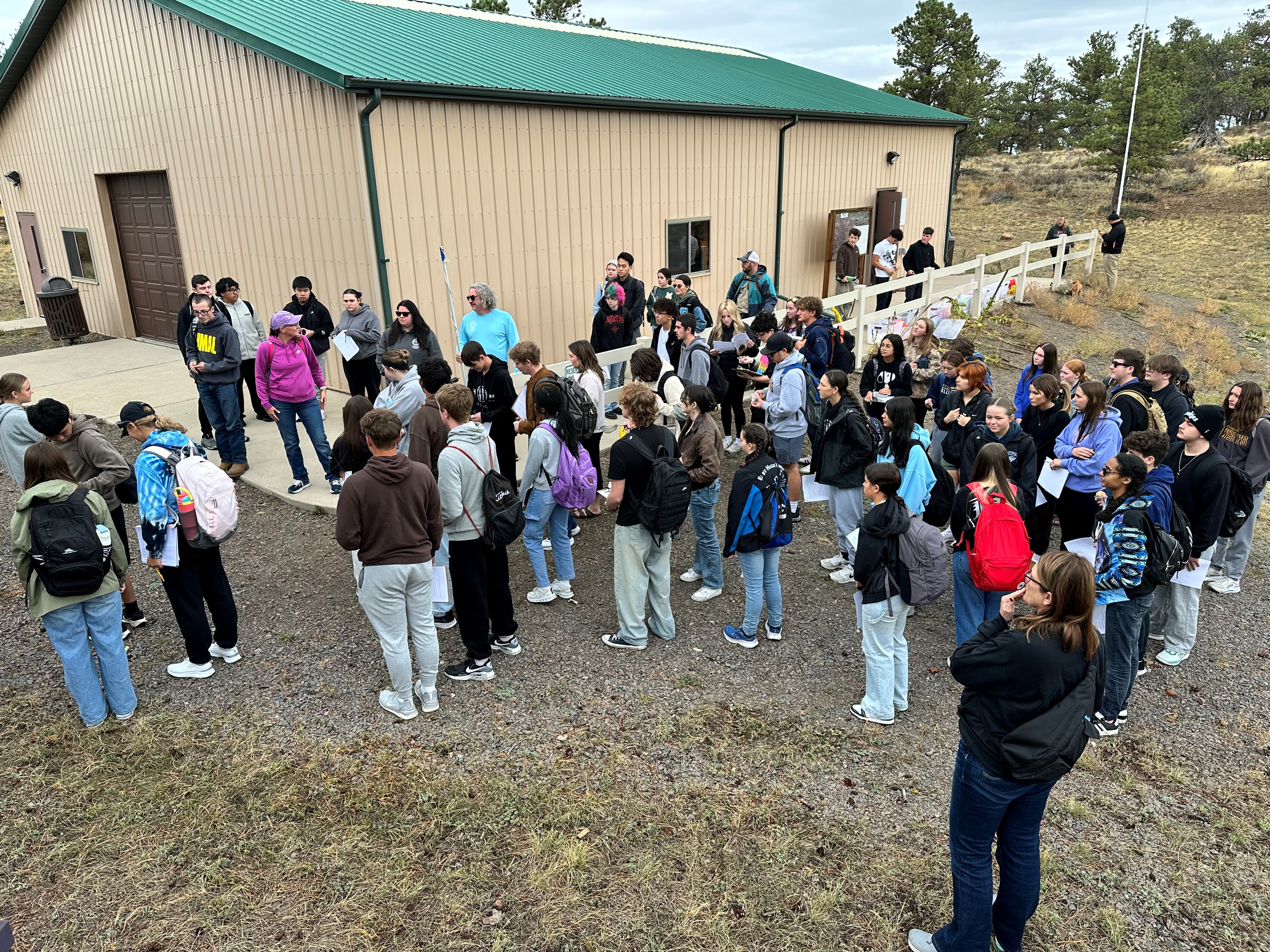A large group of students with backpacks gathers outside a beige building with a green roof.