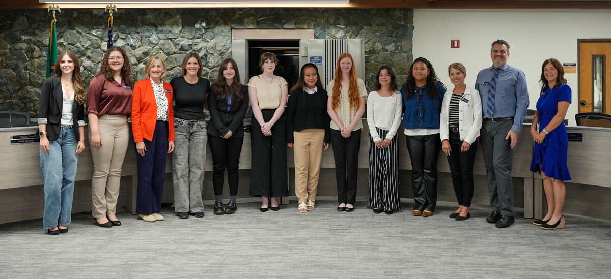 A diverse group of women and two men stand in a line in front of a stone wall and wooden paneling.