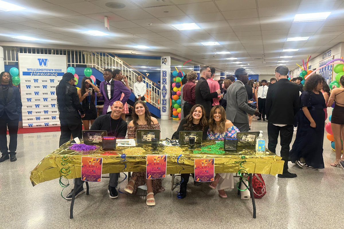 Four smiling adults sit behind a gold-draped check-in table at a Williamstown High School homecoming event.