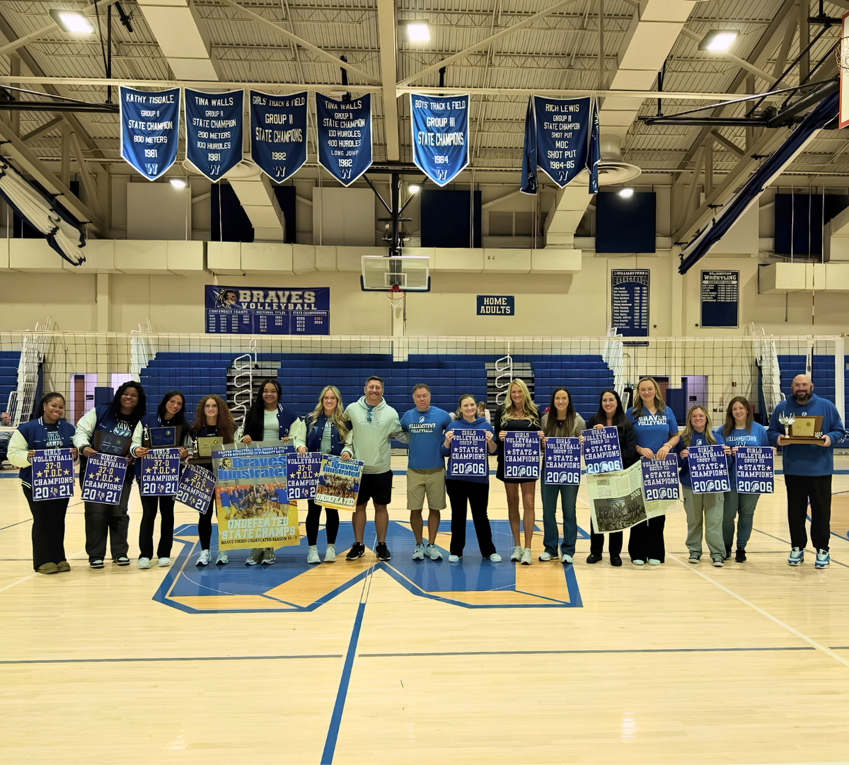 A volleyball team and coaches stand proudly in a gymnasium, holding championship banners and trophies.