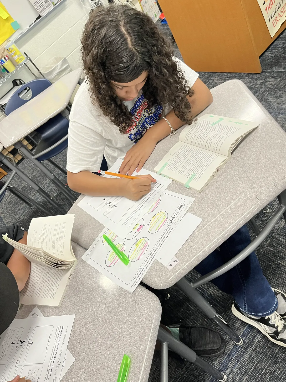 A student sits at a desk in a classroom, focused on writing in a workbook while reading an open book.