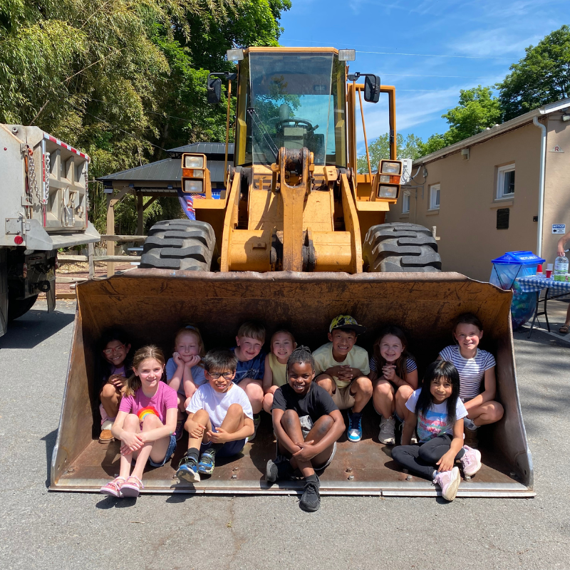 A group of smiling children sit in the bucket of a large yellow front-end loader.