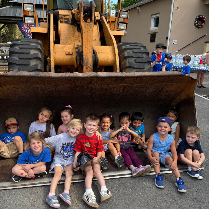 A group of children sit inside the bucket of a large yellow front-end loader.