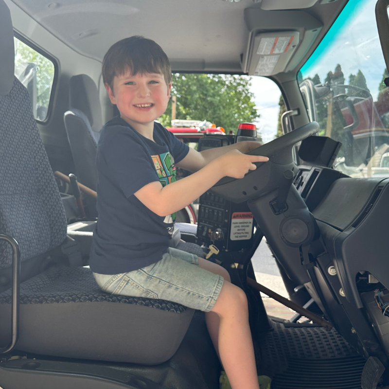 A young boy smiles while sitting in the driver's seat of a fire truck.