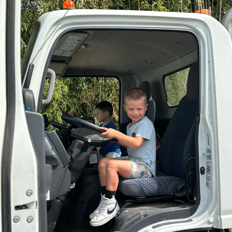 Two young boys sit in the driver's seat of a white truck, one smiling at the camera.