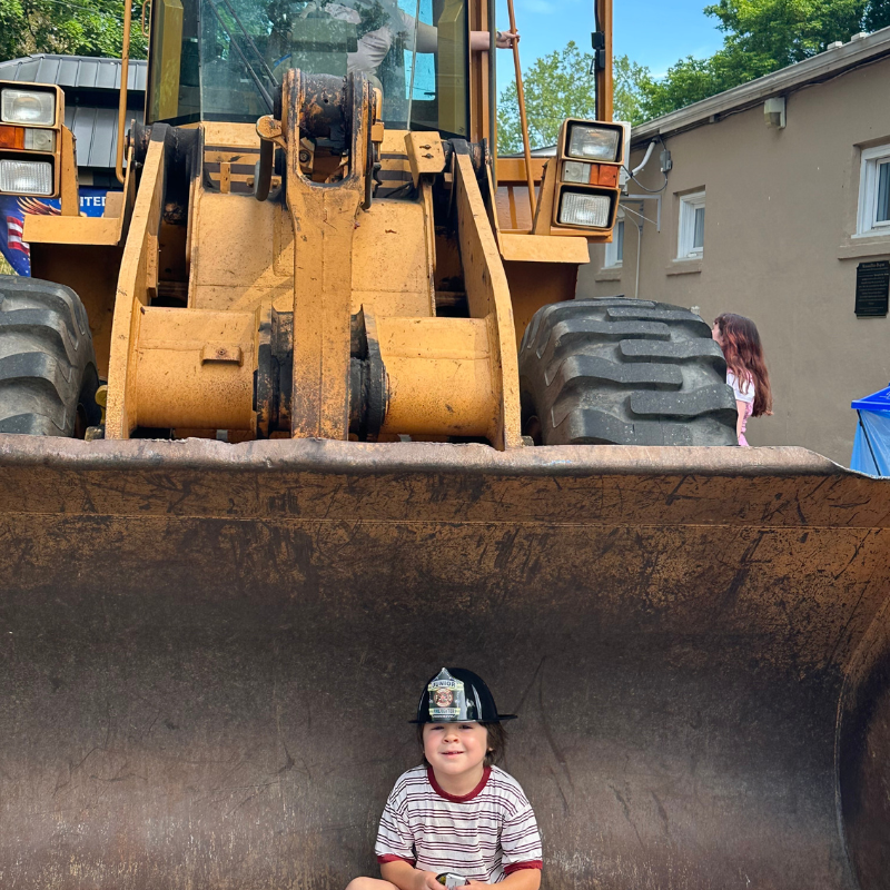 A young boy wearing a junior firefighter helmet sits inside the bucket of a large yellow front-end loader.
