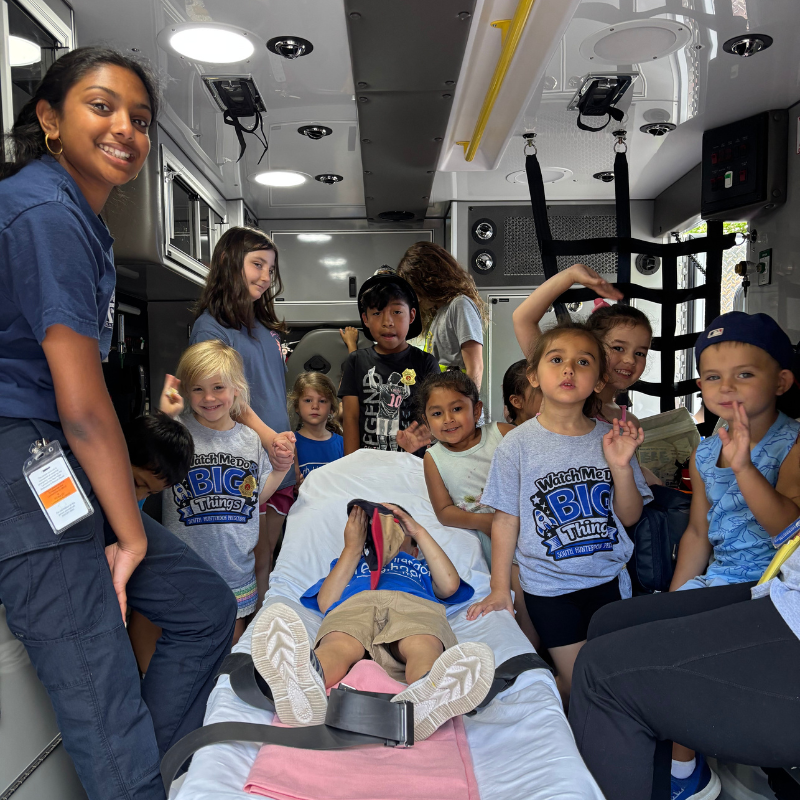 A group of children and an adult stand inside an ambulance, with one child lying on a stretcher.