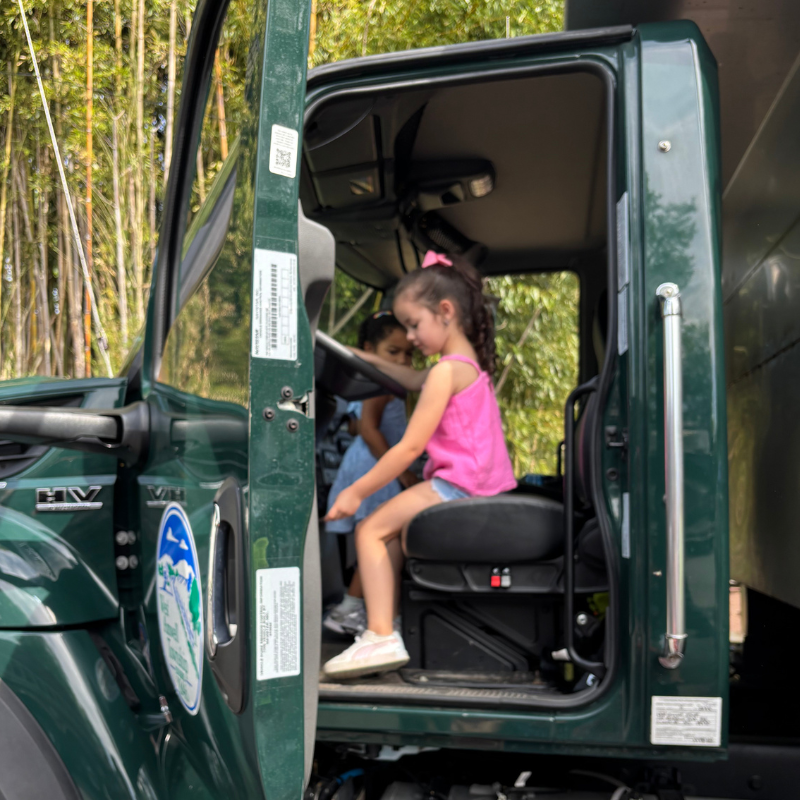 Two young girls sit in the driver's seat of a green truck, with one in the foreground wearing a pink top.