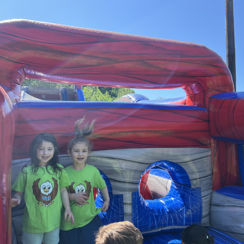 Two smiling children in matching green t-shirts stand inside a large, colorful inflatable bounce house.