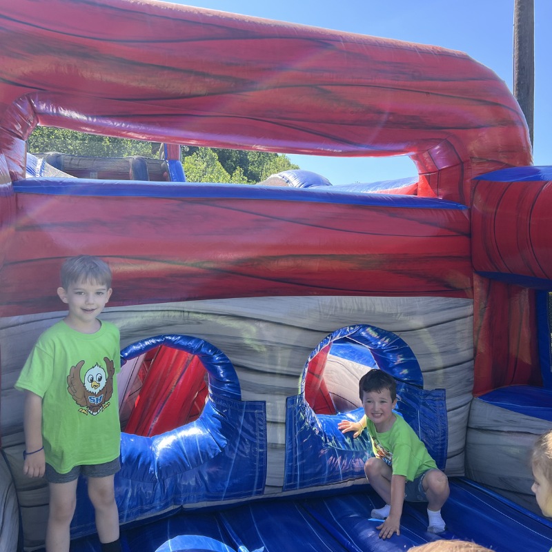 Two young boys in green t-shirts smile inside a large, colorful inflatable bounce house.