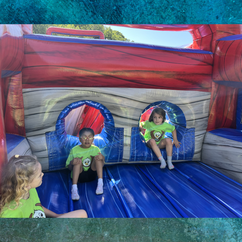 Three children in matching lime green shirts with owl graphics are playing inside a large inflatable bounce house.