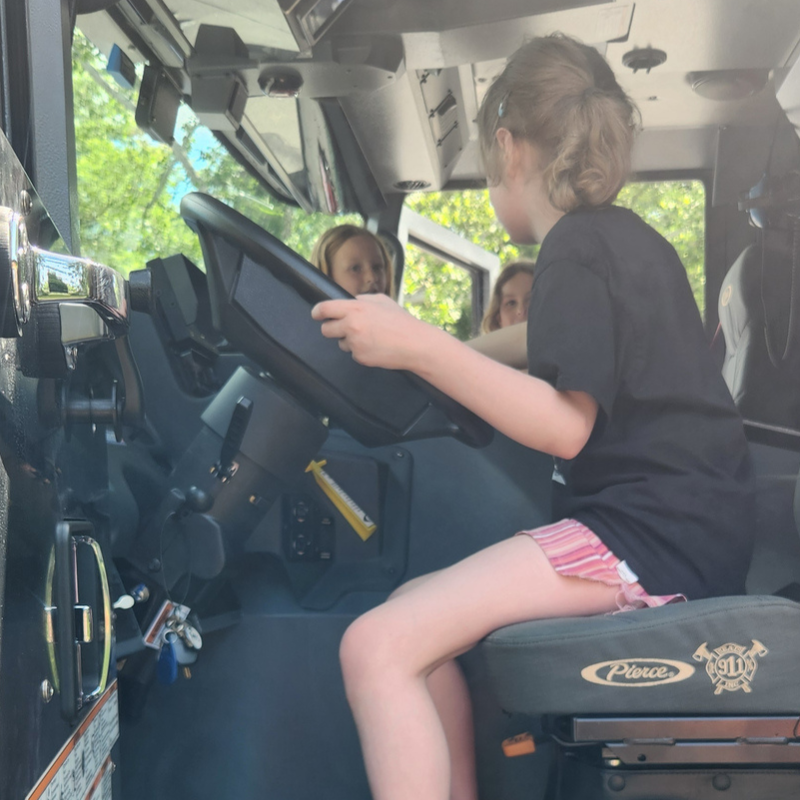 A young girl sits in the driver's seat of a fire truck, holding the steering wheel.