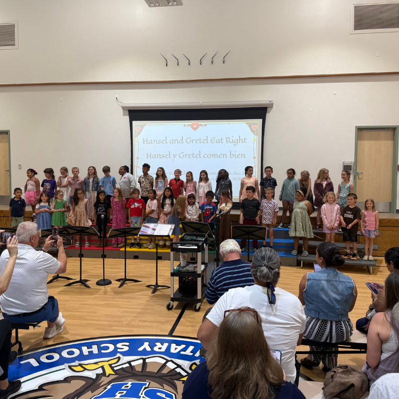 A group of children stand on risers on a stage, performing in front of an audience.