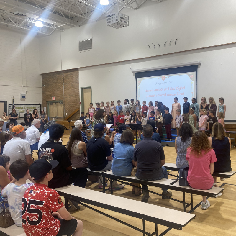A group of children stand on a stage in a gymnasium, performing a song titled 'Hansel and Gretel Eat Right!'