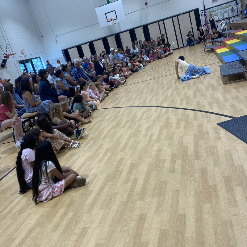 An audience of children and adults sit on a gymnasium floor watching a performer.