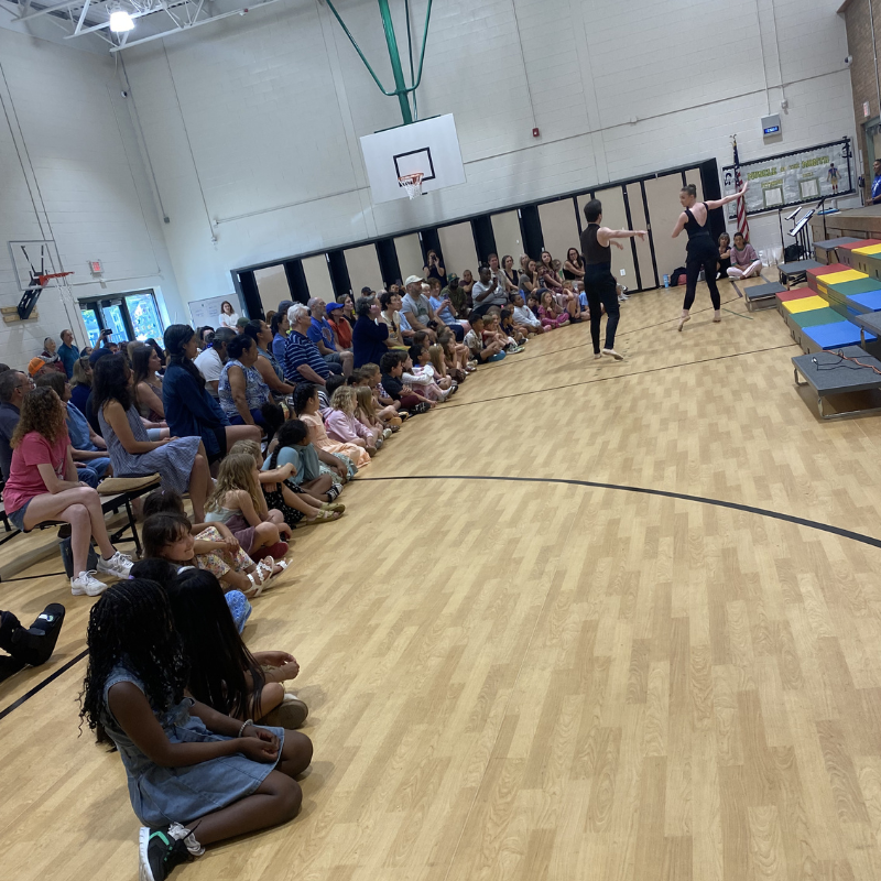 An audience of children and adults sit on a gymnasium floor watching a dance performance.