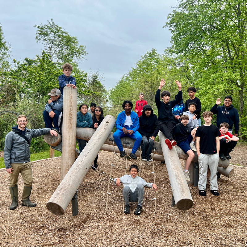 A group of children and an adult pose on a wooden playground structure surrounded by trees.
