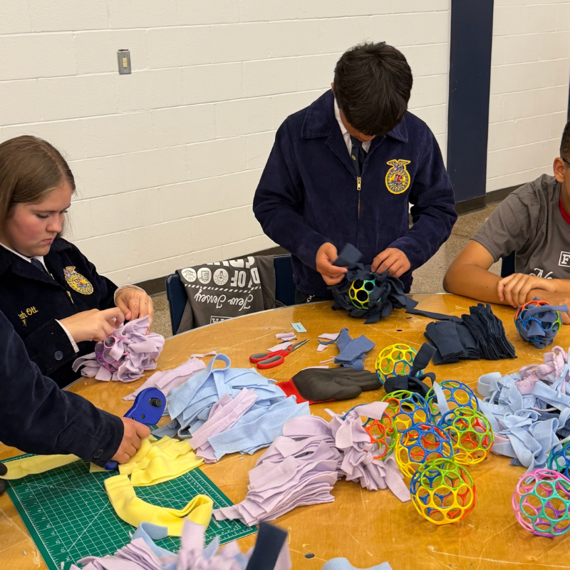 Students in FFA jackets work together at a table, crafting colorful fabric balls.