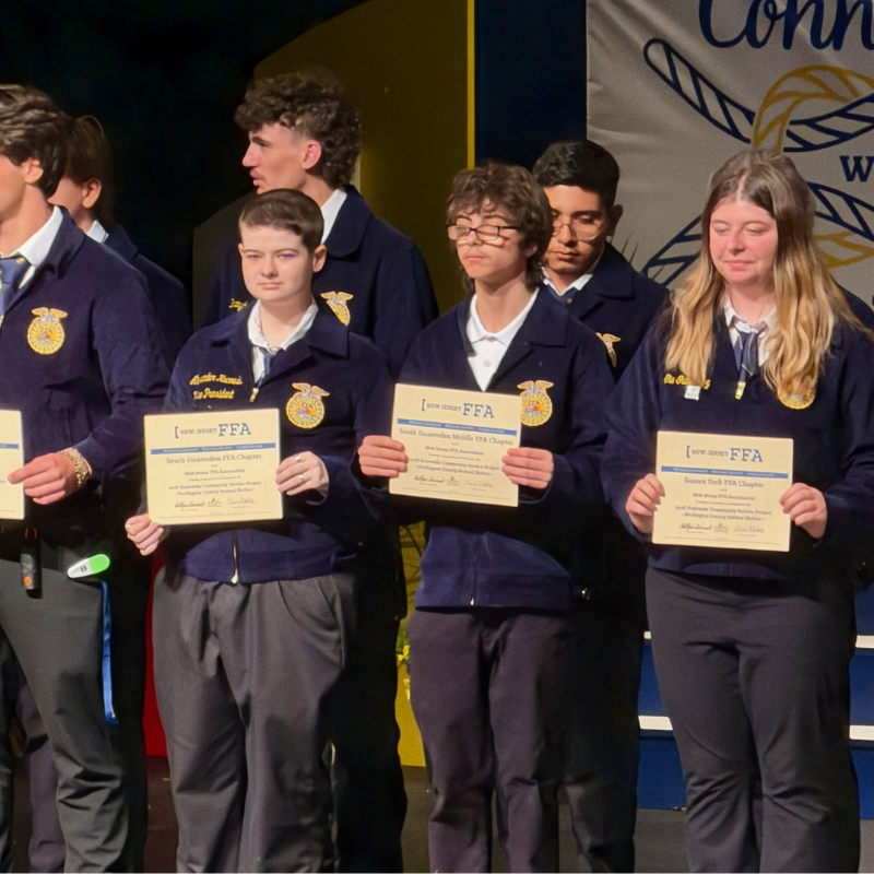 Students in FFA jackets stand on a stage holding certificates, with "New Jersey FFA" visible on the awards.