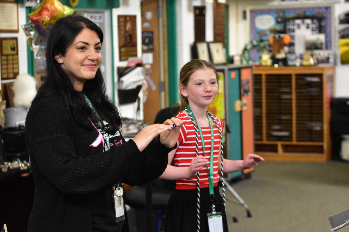 A woman and a young girl stand in a classroom, both with their hands raised as if demonstrating a movement.