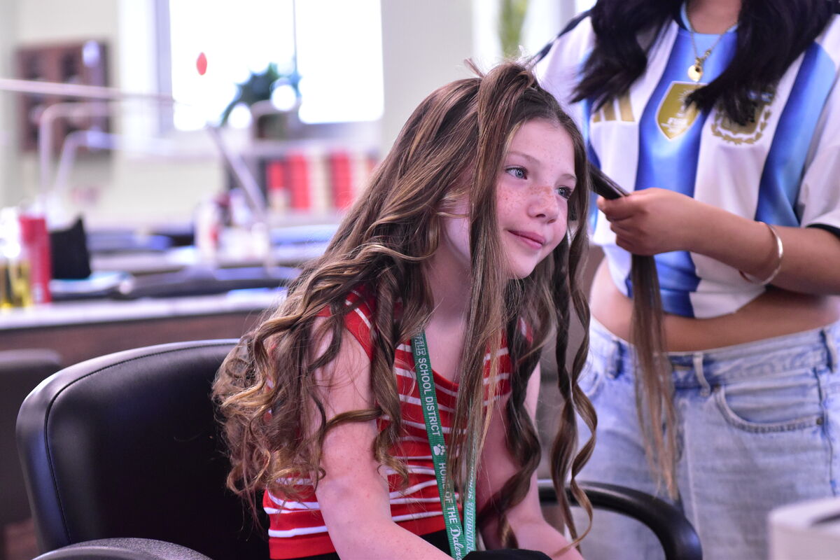 A young girl with freckles and long, wavy brown hair smiles as someone styles her hair.
