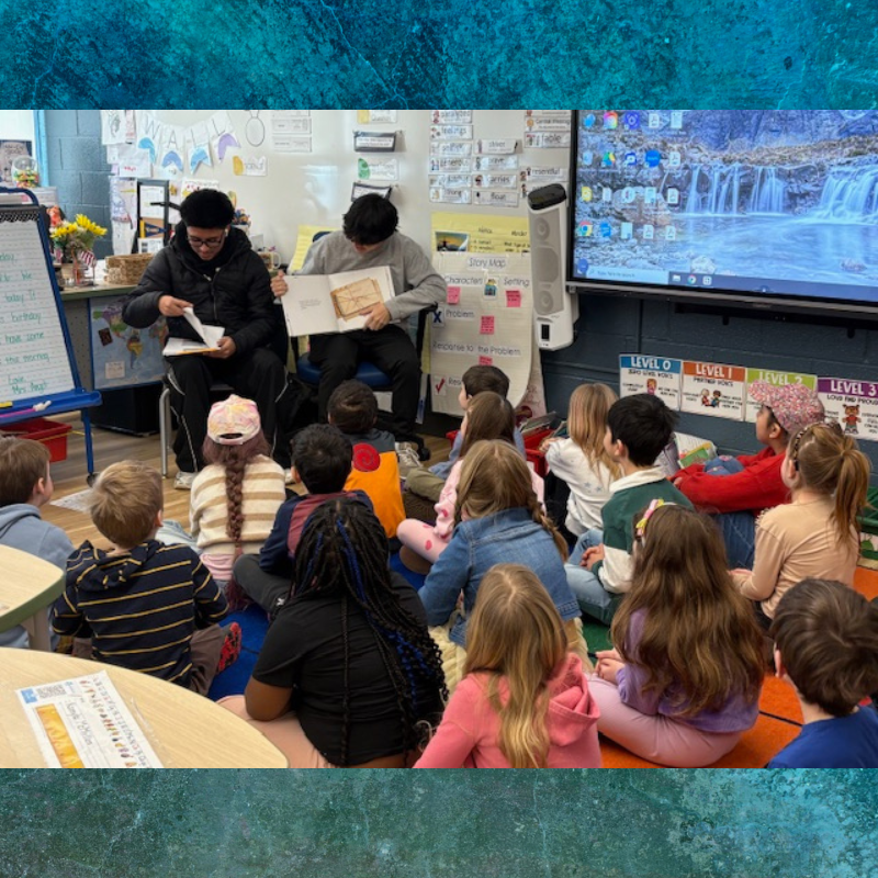 Two students read a book aloud to a class of young children sitting on the floor.
