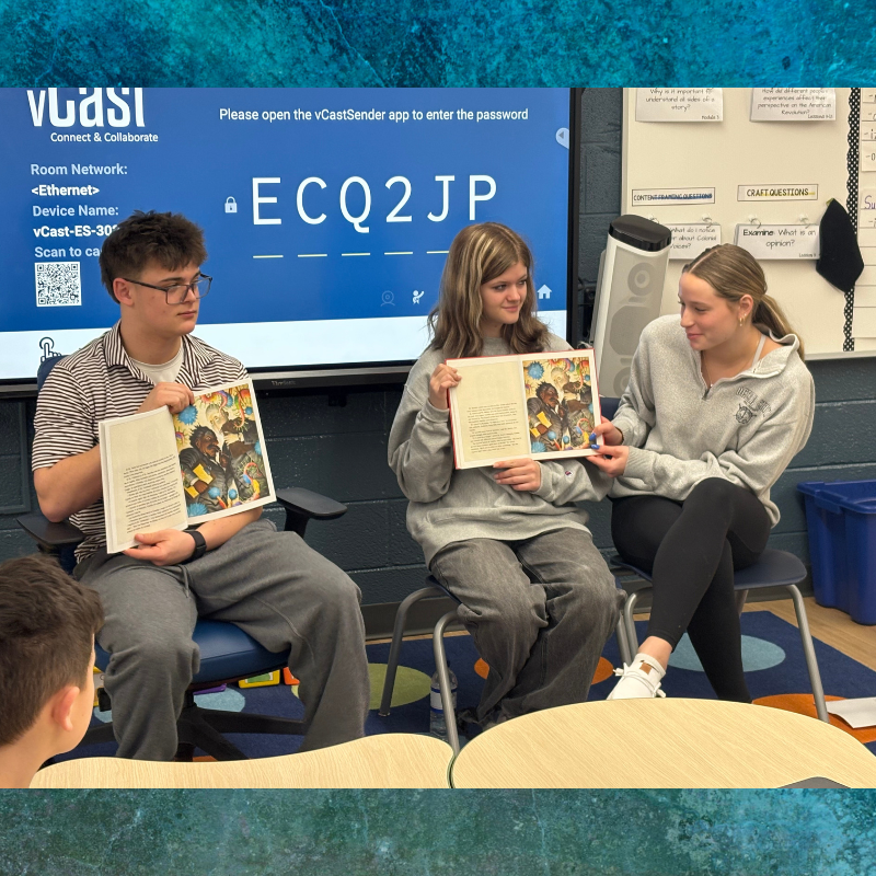 Three students sit in a classroom, holding open a book with colorful illustrations.
