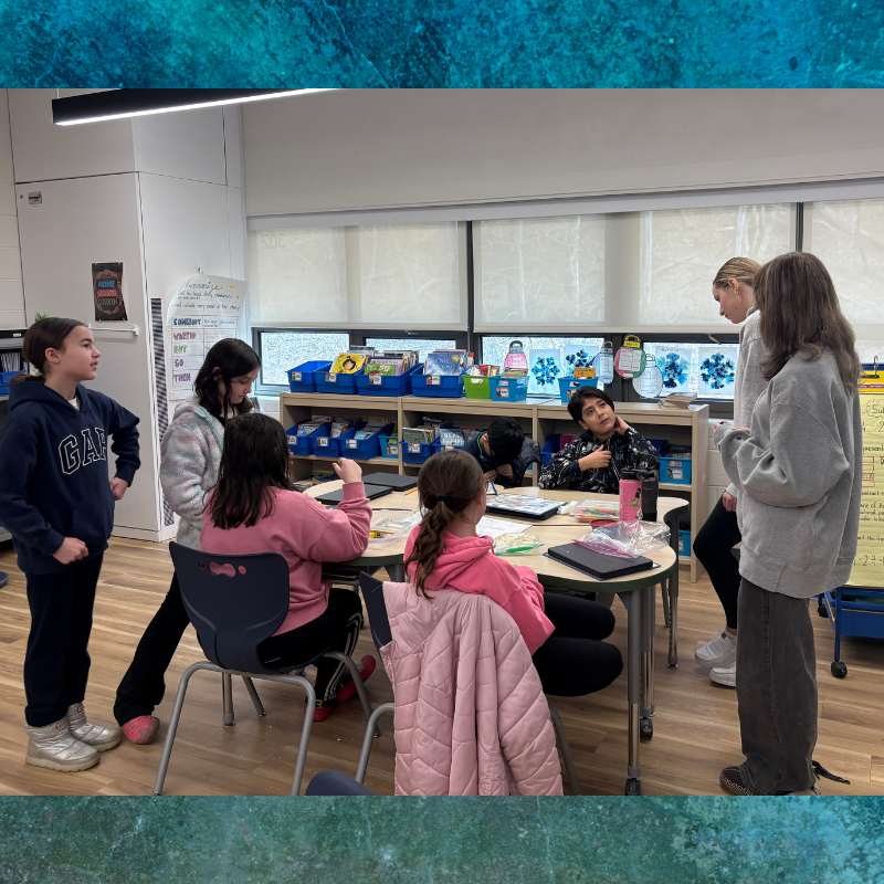 Students collaborate around a table in a classroom, discussing a lesson.