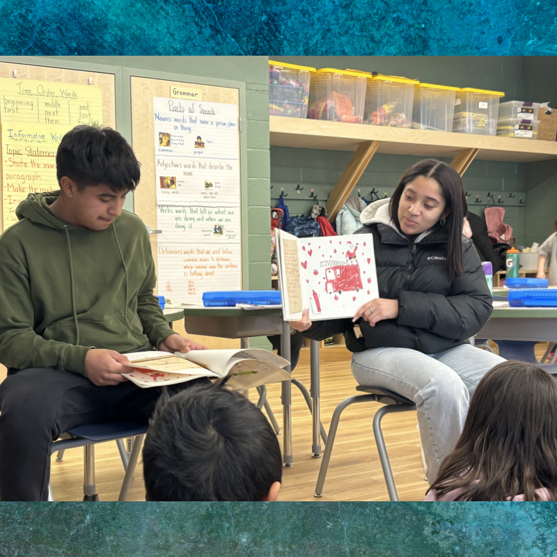 A young woman reads a book with colorful illustrations to a group of students in a classroom.