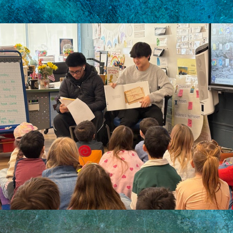 Two young men read a book to a group of children sitting on the floor in a classroom.