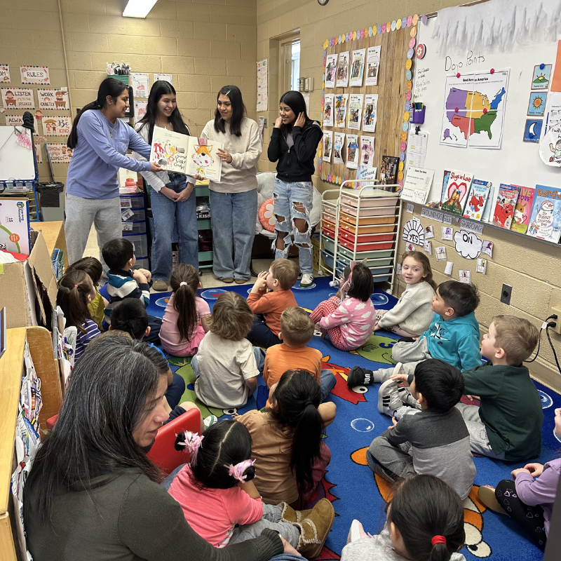 Four young women read a book to a group of children sitting on a colorful rug in a classroom.